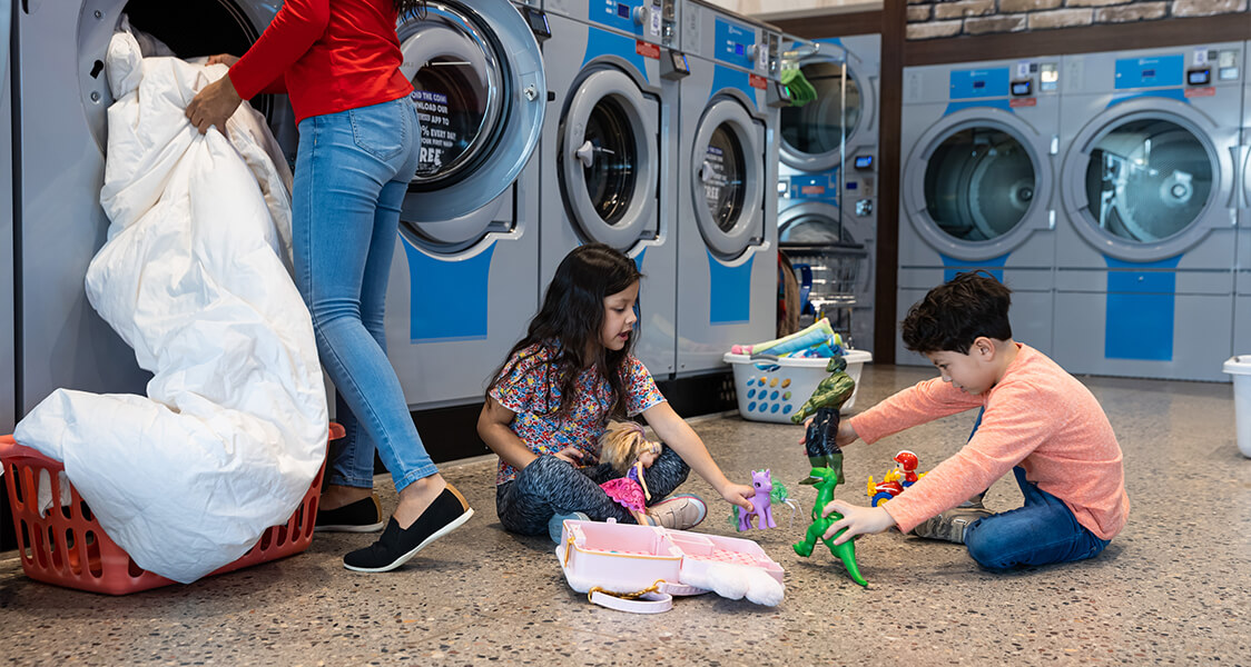 two kids playing with toys on the floor as their mom does the laundry